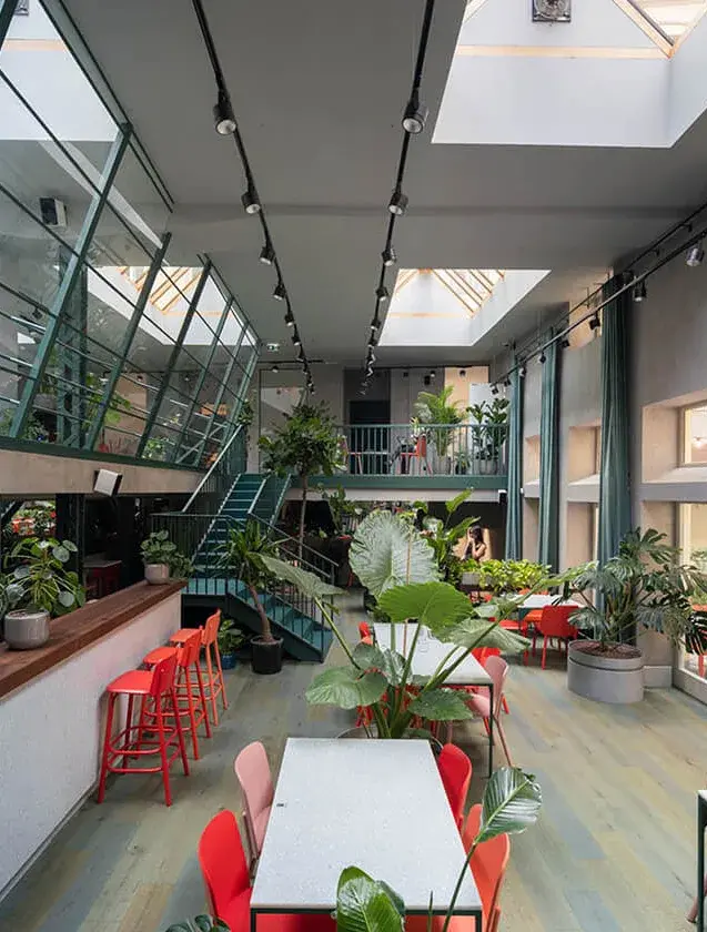 Celkový pohled na víceúrovňový interiér Healthy Longevity Café s bílými stoly a barevnými židlemi, kterému dominuje ocelové schodiště a prosklená šikmá stěna propojující patra.