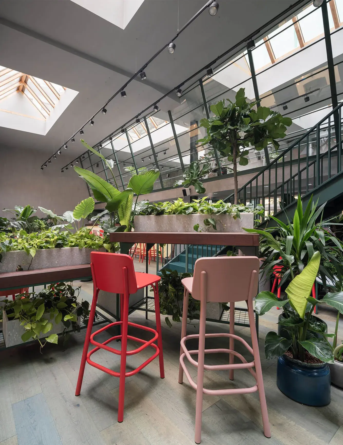 Interiér kavárny Healthy Longevity Café s detailem červené a růžové barové židle u dřevěného pultu, obklopený bohatou vegetací a v pozadí se zeleným schodištěm vedoucím do patra pod prosklenou střechou.