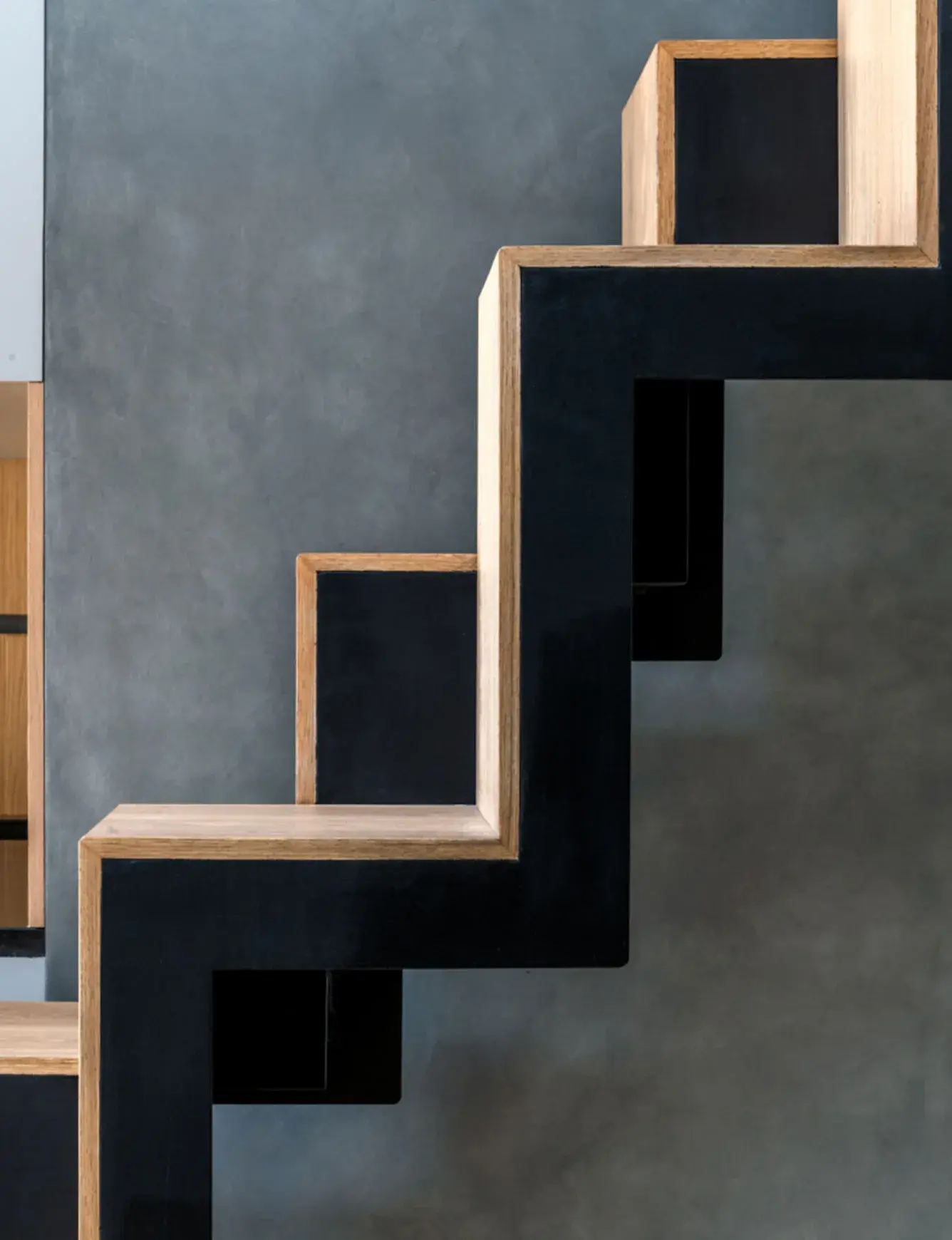 Detailní pohled na geometrickou konstrukci černého „zubatého“ schodiště s nášlapy z přírodního dubu, které kontrastuje s tmavě šedou stěnou s betonovou stěrkou v interiéru Karlínského útočiště.