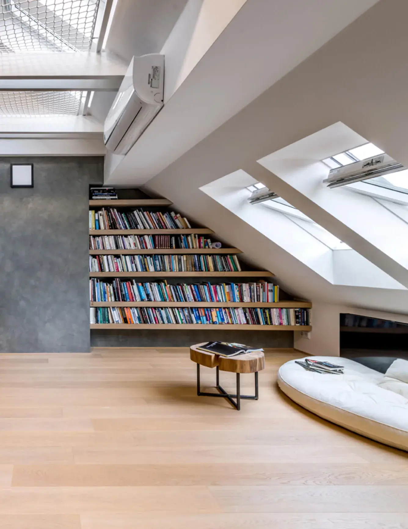 Detailní pohled na atypickou vestavěnou knihovnu v podkroví bytu Karlínské útočiště, jejíž police kopírují sklon střechy vedle střešních oken, doplněnou o dubovou podlahu a relaxační zónu s kulatým sedacím vakem.