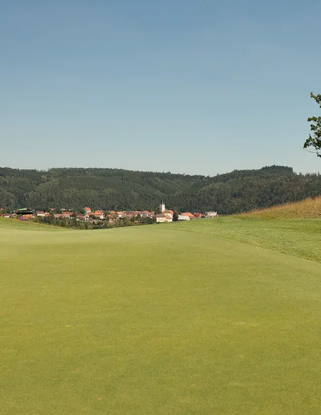 Panoramatický pohled na rozlehlé zelené golfové hřiště v Kácově, v jehož pozadí se rozkládá obec s kostelní věží obklopená zalesněnými kopci Posázaví.