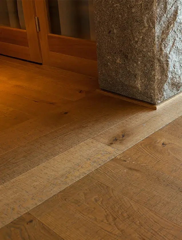 Detailní pohled na precizní zakončení rustikální dubové podlahy u paty masivního kamenného sloupu v Panorama Golf Resortu, zvýrazňující texturu kartáčovaného dřeva a přírodní kresbu lamel.