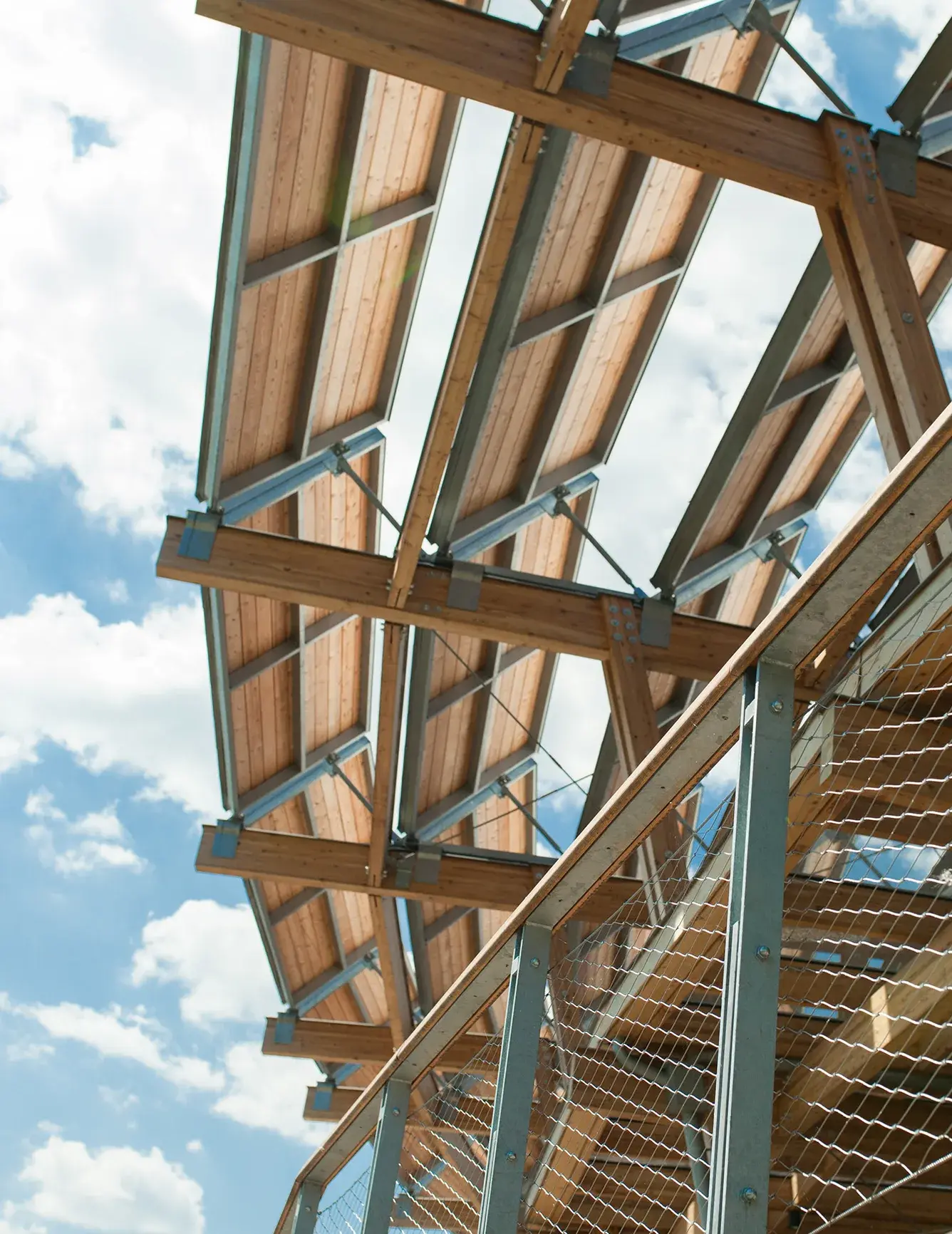 Podhled na unikátní dřevěnou konstrukci střechy Panorama Golf Resortu v Kácově od architekta Martina Rajniše, kombinující masivní trámy, dřevěné panely a ocelové prvky proti modré obloze.