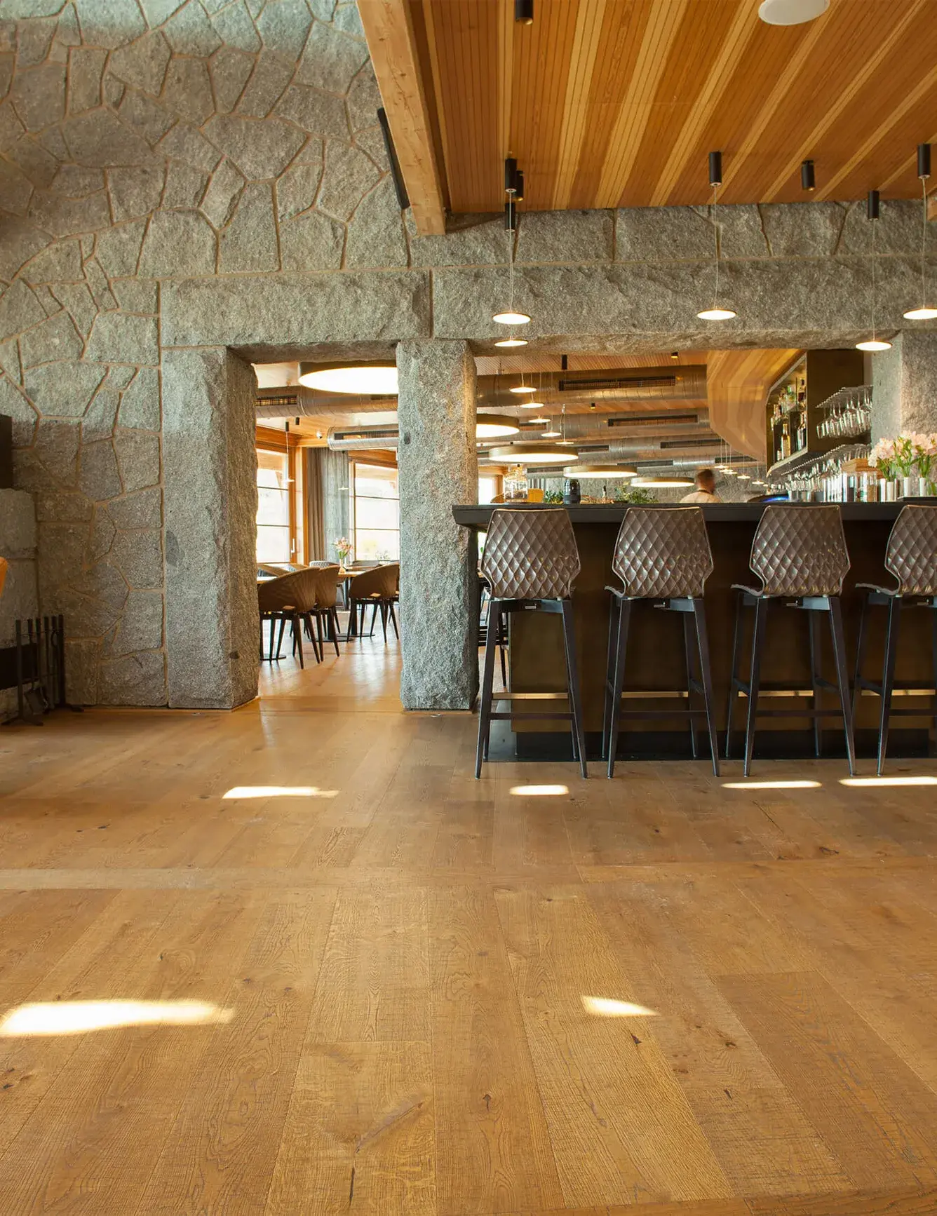 Prosvětlený interiér restaurace a baru v Panorama Golf Resortu s rustikální dubovou podlahou, kterou doplňují masivní kamenné zdi, prošívané barové židle a moderní lineární osvětlení nad barem.