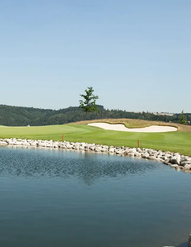 Pohled na vodní plochu a travnatý green v Panorama Golf Resortu, s písečným bunkrem v pozadí a zalesněnými kopci Posázaví na obzoru.