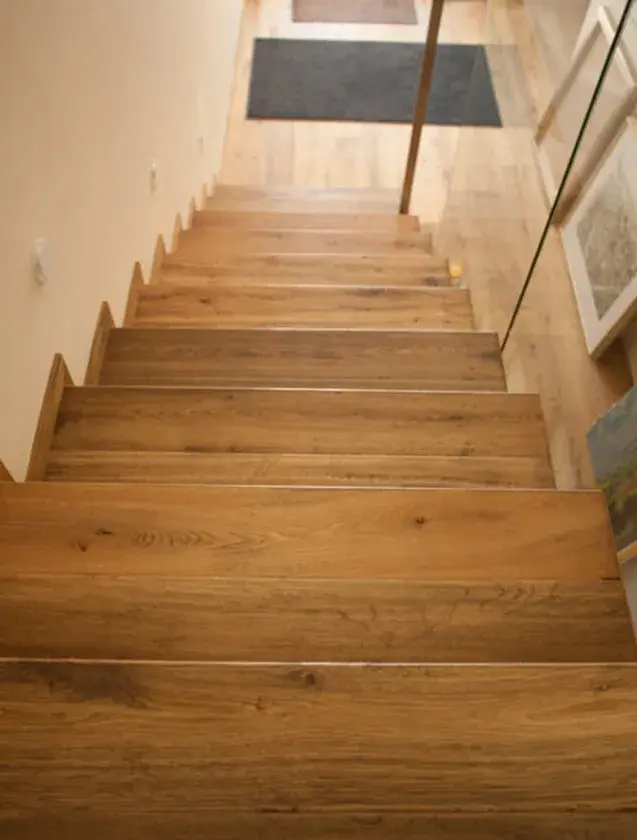Pohled z horní části schodiště v domě v Uherském Hradišti, kde dubové stupně a navazující podlaha z kolekce Oakcent Honey s ručně sraženými hranami a Nano olejem vytvářejí hřejivou, medovou atmosféru v kombinaci s moderním skleněným zábradlím.
