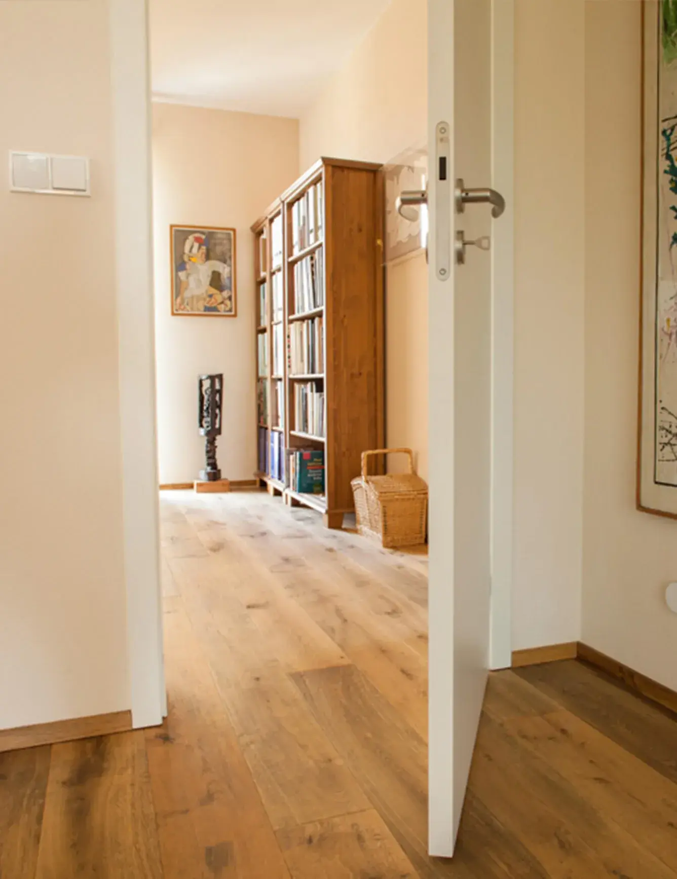 Pohled otevřenými dveřmi do světlé chodby rodinného domu v Uherském Hradišti, kde dubová podlaha Oakcent Rustic s nano olejem a běleným, louhovaným povrchem tvoří harmonický celek s dřevěnou knihovnou a minimalistickým bílým interiérem.