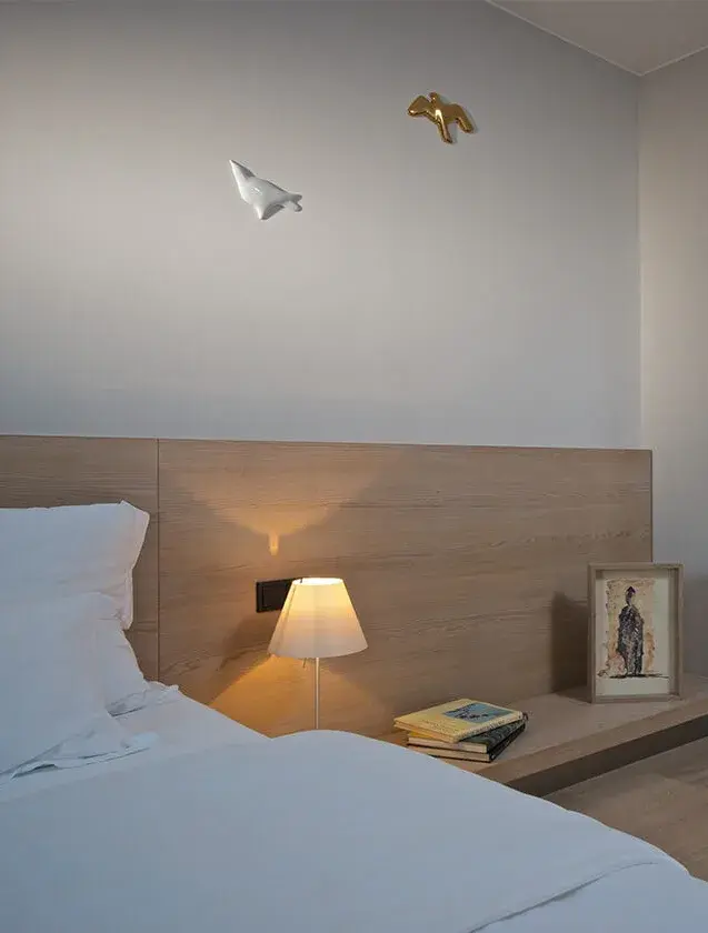 Minimalistický detail ložnice v pražském apartmánu s dřevěným obložením stěny u lůžka, noční lampičkou a dekorativními prvky na šedé stěně, doplňující nadčasový design kolekce Sand.