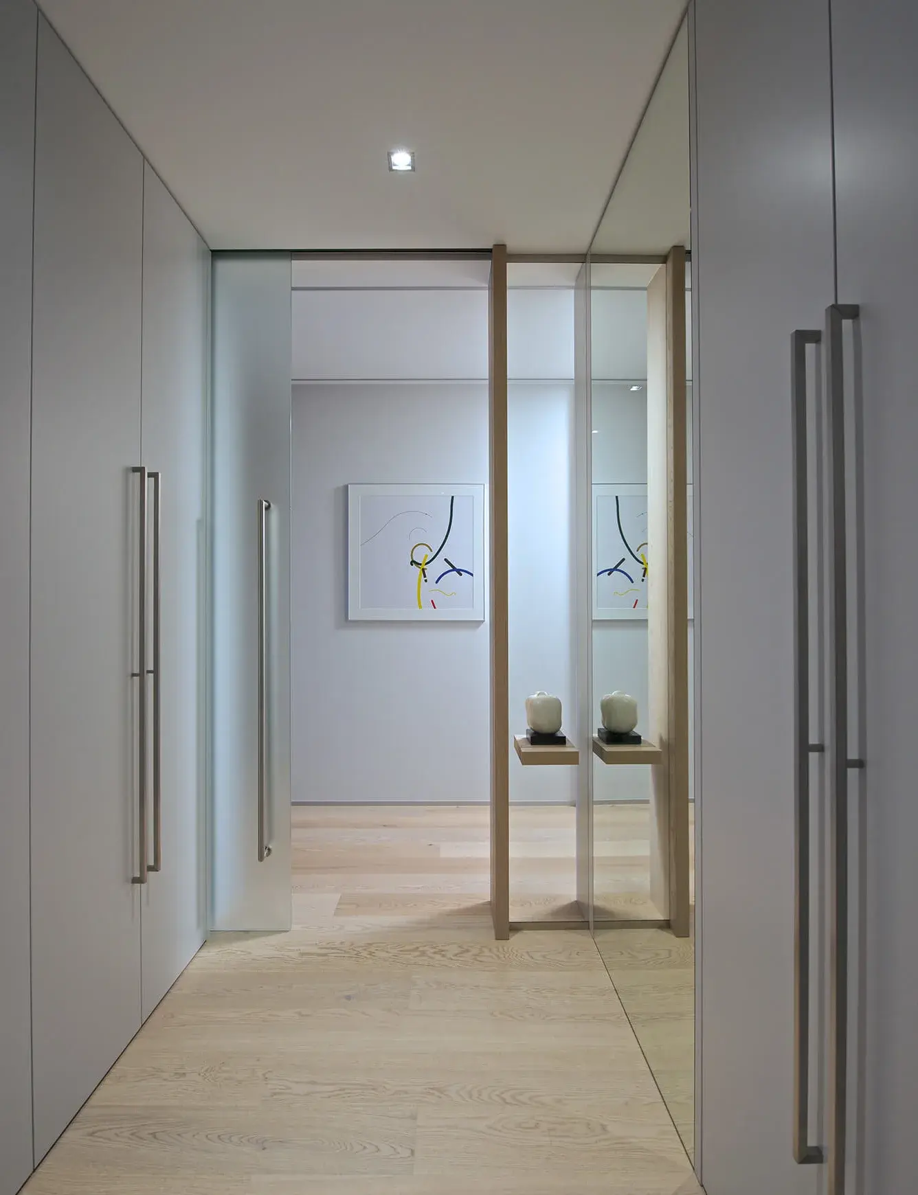 Minimalistická chodba pražského apartmánu s dubovou podlahou Oakcent Rustic v dekoru Planks, kde světlý odstín dřeva Milky doplňují bílé vestavné skříně s nerezovými madly a velké zrcadlo, které opticky rozšiřuje prostor.