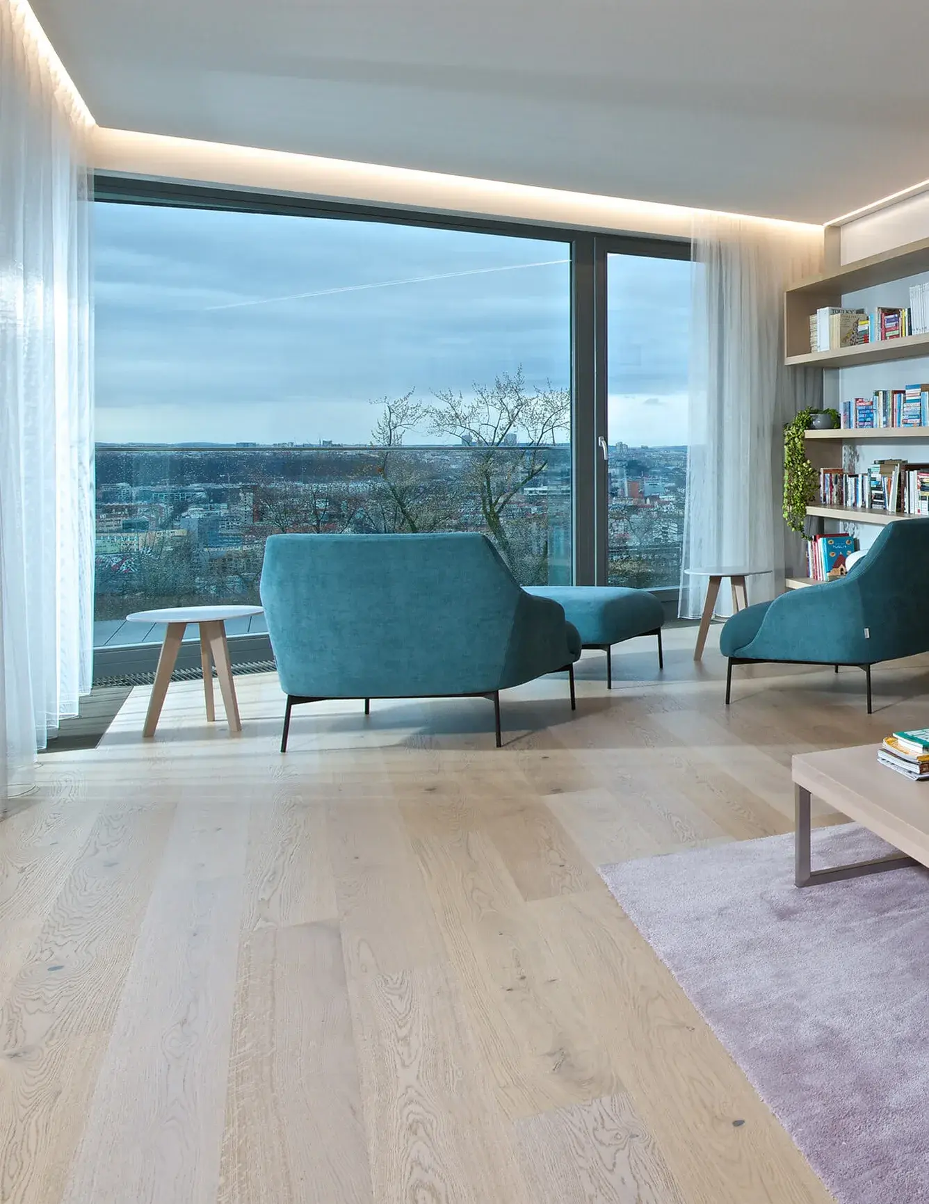 Prosvětlený obývací pokoj v pražském apartmánu s dubovou podlahou Oakcent v odstínu Milky, kterému dominují dvě tyrkysová designová křesla a velkoformátové okno s panoramatickým výhledem na Prahu.