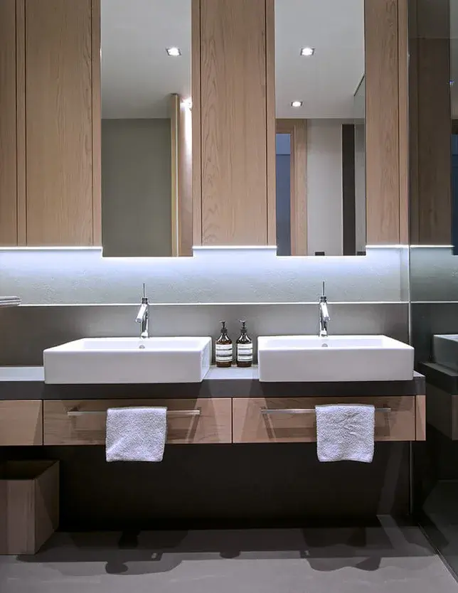 Moderní koupelna v pražském apartmánu s dvojumyvadlem na zavěšené dubové skříňce, kterou doplňuje velké zrcadlo s integrovaným LED podsvícením a dřevěné obklady stěn ladící s podlahou Oakcent v odstínu Milky.