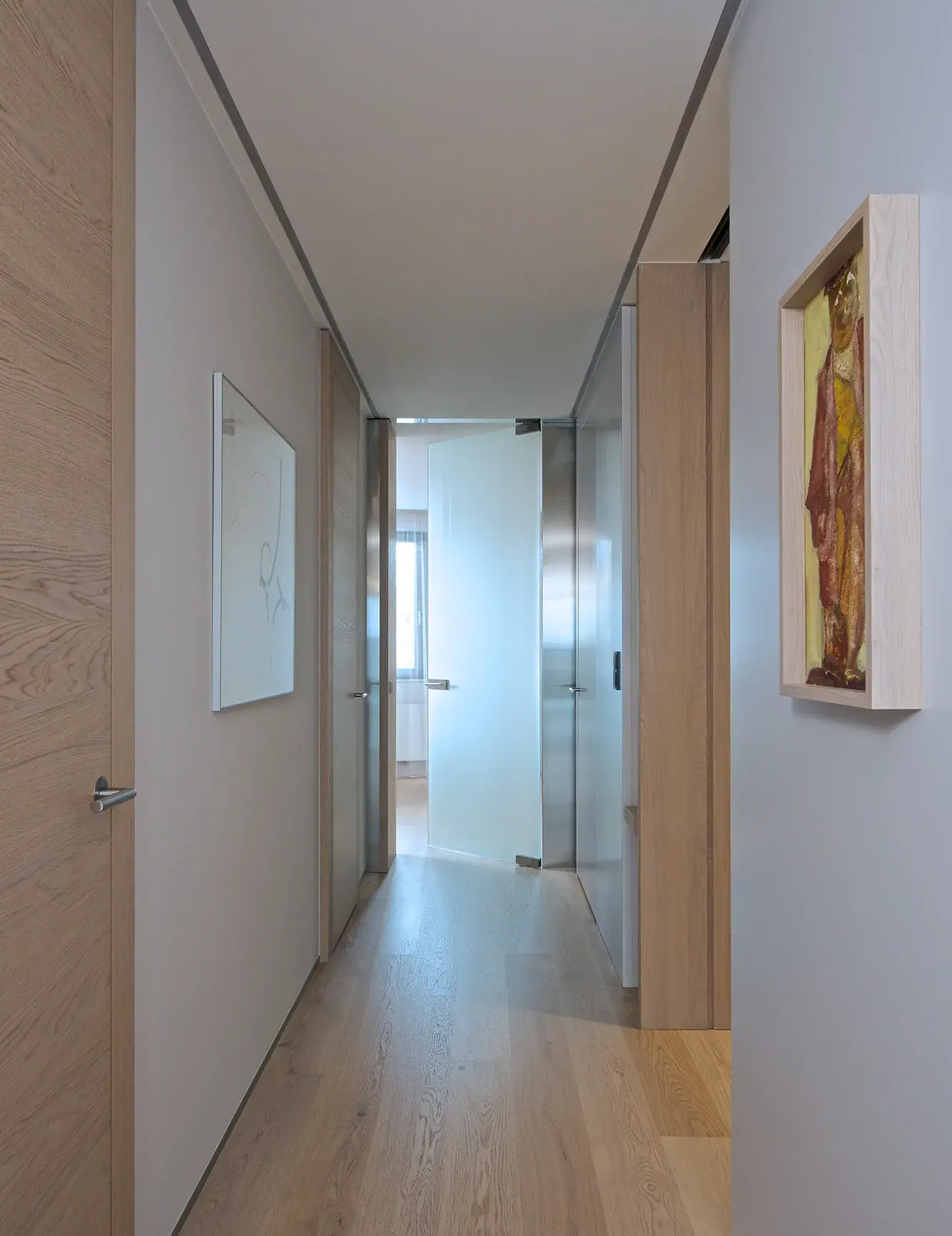 Pohled do minimalistické chodby pražského apartmánu s dubovou podlahou Oakcent Rustic Milky, lemovanou světlými stěnami s uměleckými obrazy a dřevěným obložením, vedoucí k proskleným dveřím.