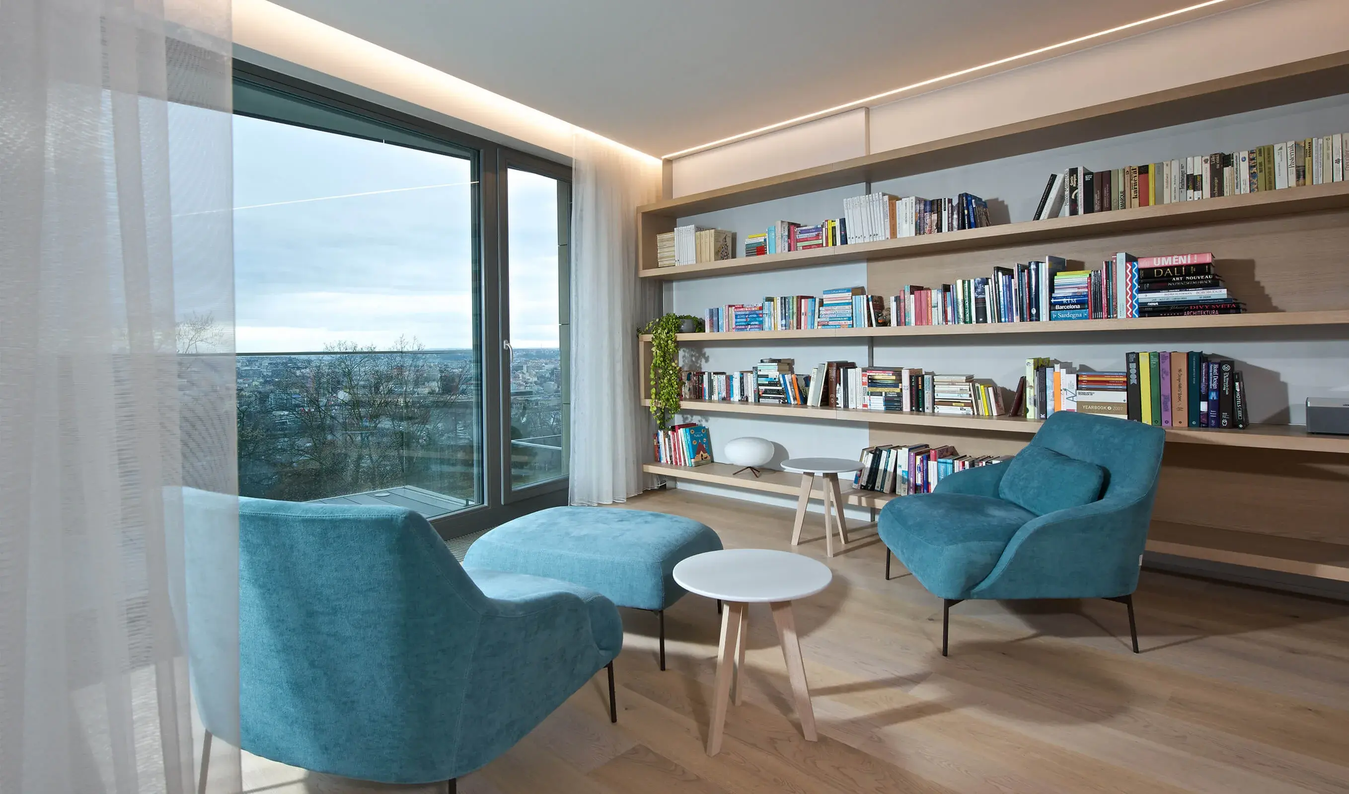 Moderní obývací pokoj v pražském apartmánu s celostěnnou dřevěnou knihovnou a dubovou podlahou Oakcent Rustic Milky, doplněný o dvě tyrkysová křesla s taburetem a velkorysé prosklení s výhledem na město.