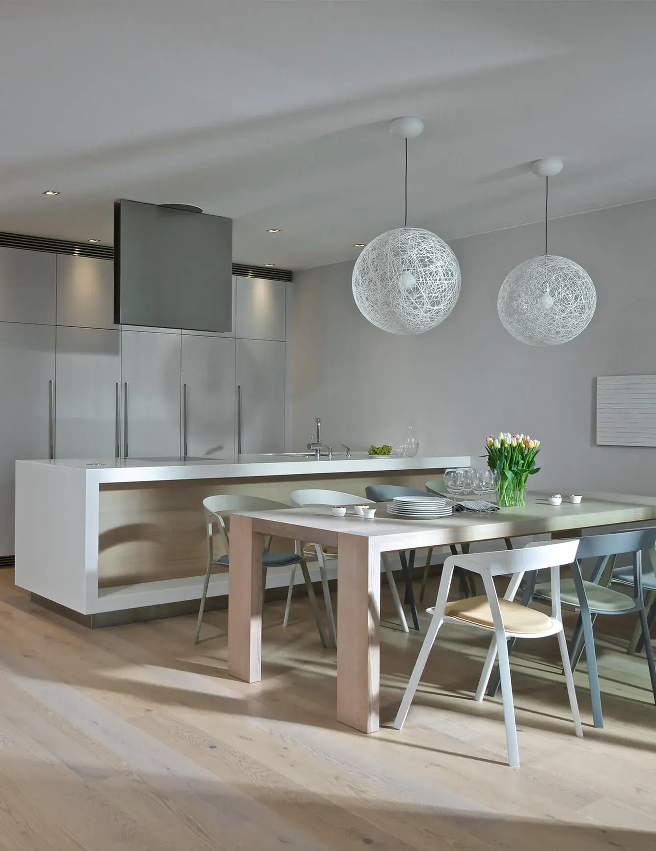 Prosvětlený interiér pražského apartmánu s jídelnou a minimalistickou kuchyní, kde dubovou podlahu Oakcent Rustic Milky doplňuje moderní bílý kuchyňský ostrůvek, dřevěný jídelní stůl a dvě výrazná kulatá svítidla se síťovanou strukturou.