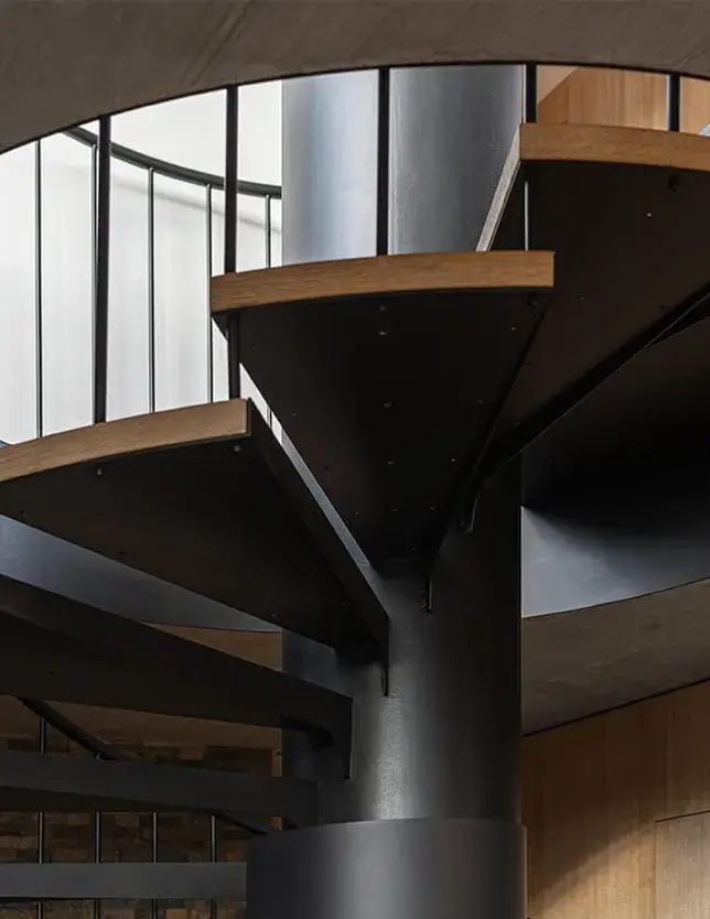 Detail černého točitého schodiště s dubovými nášlapy v interiéru Domu nad rybníkem, zdůrazňující kontrast mezi kovovou konstrukcí a hřejivou texturou dřeva.