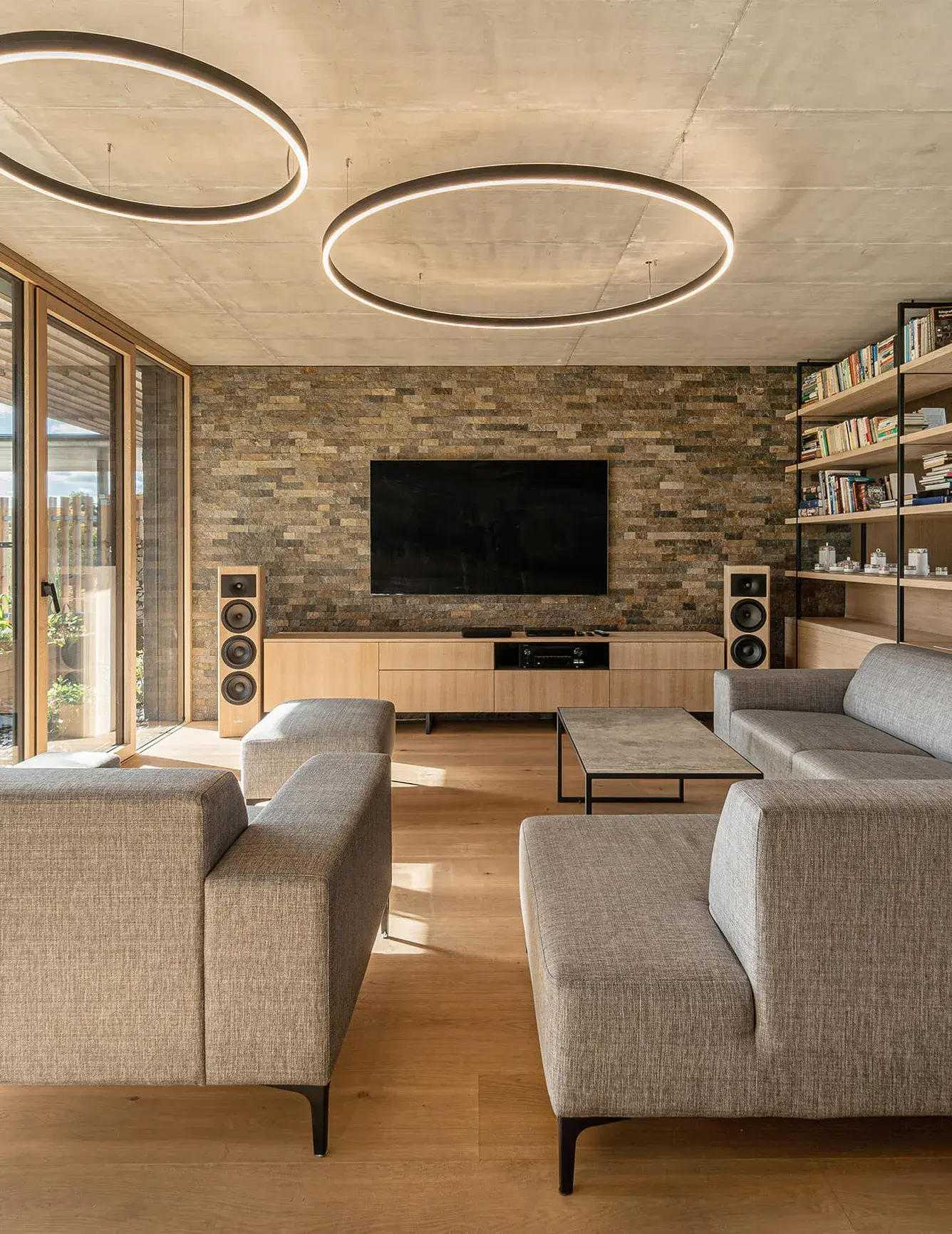 Pohled na obývací pokoj v Domu nad rybníkem s šedou pohovkou a křesly, kde kamenný obklad stěny s televizí doplňuje dubová podlaha a moderní kruhová svítidla na betonovém stropě.