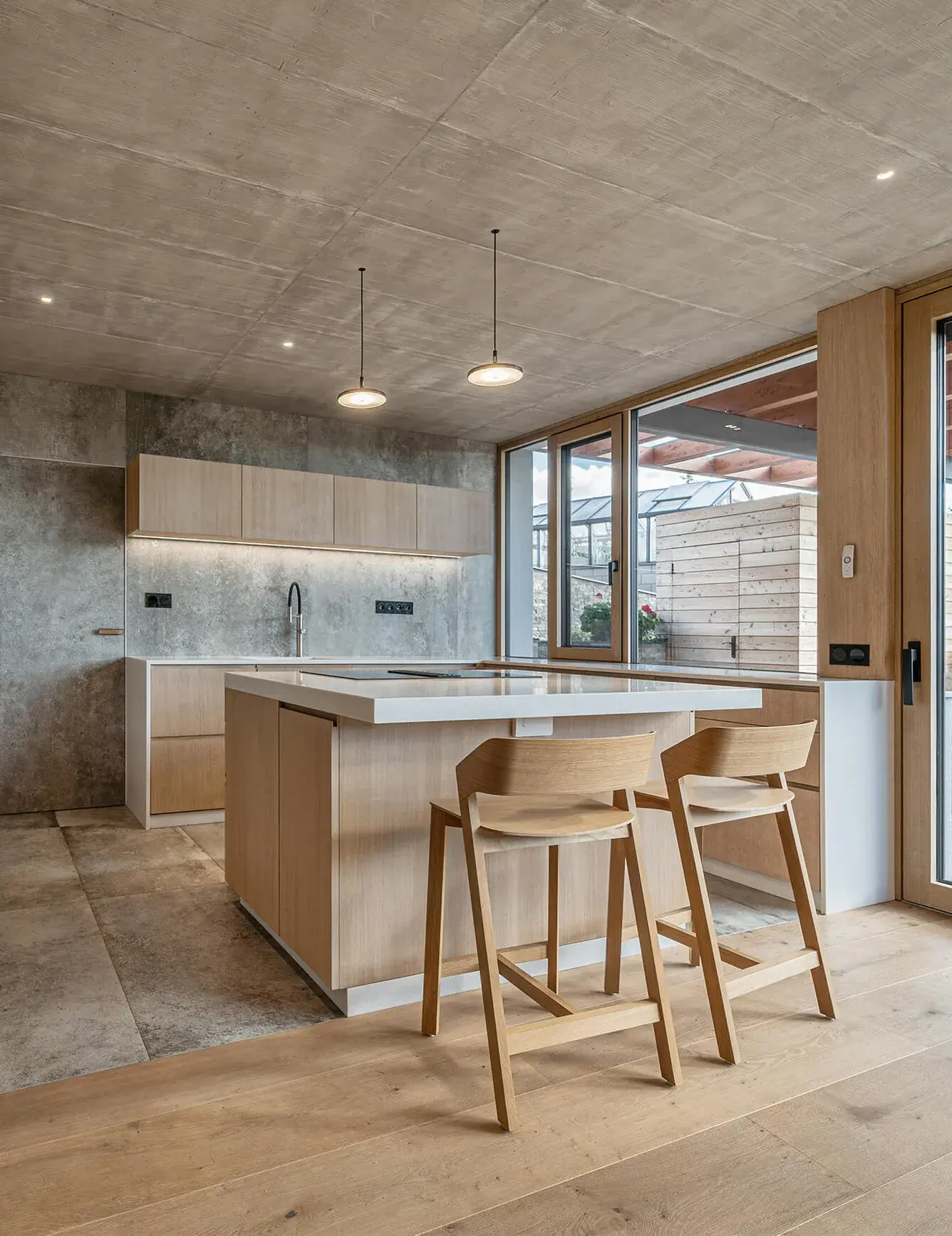 Interiér moderní kuchyně s ostrůvkem v Domu nad rybníkem, kde se snoubí pohledový betonový strop a stěrky s hřejivým dubem na podlaze, barových židlích i okenních rámech.