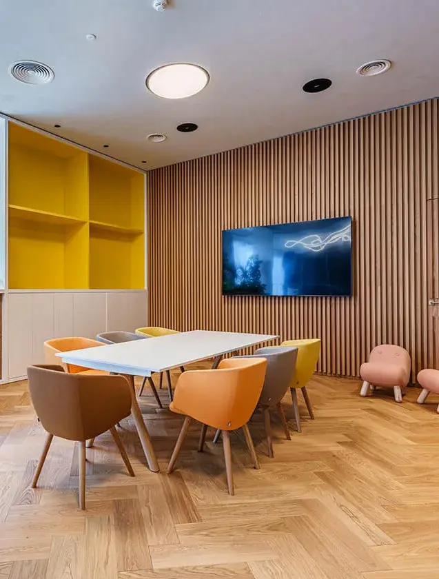 Interiér konferenční místnosti v hotelu Holiday Inn s moderním dřevěným lamelovým obkladem stěny, žlutým úložným výklenkem a barevnými křesílky kolem bílého jednacího stolu na dubové podlaze vzoru rybí kost.
