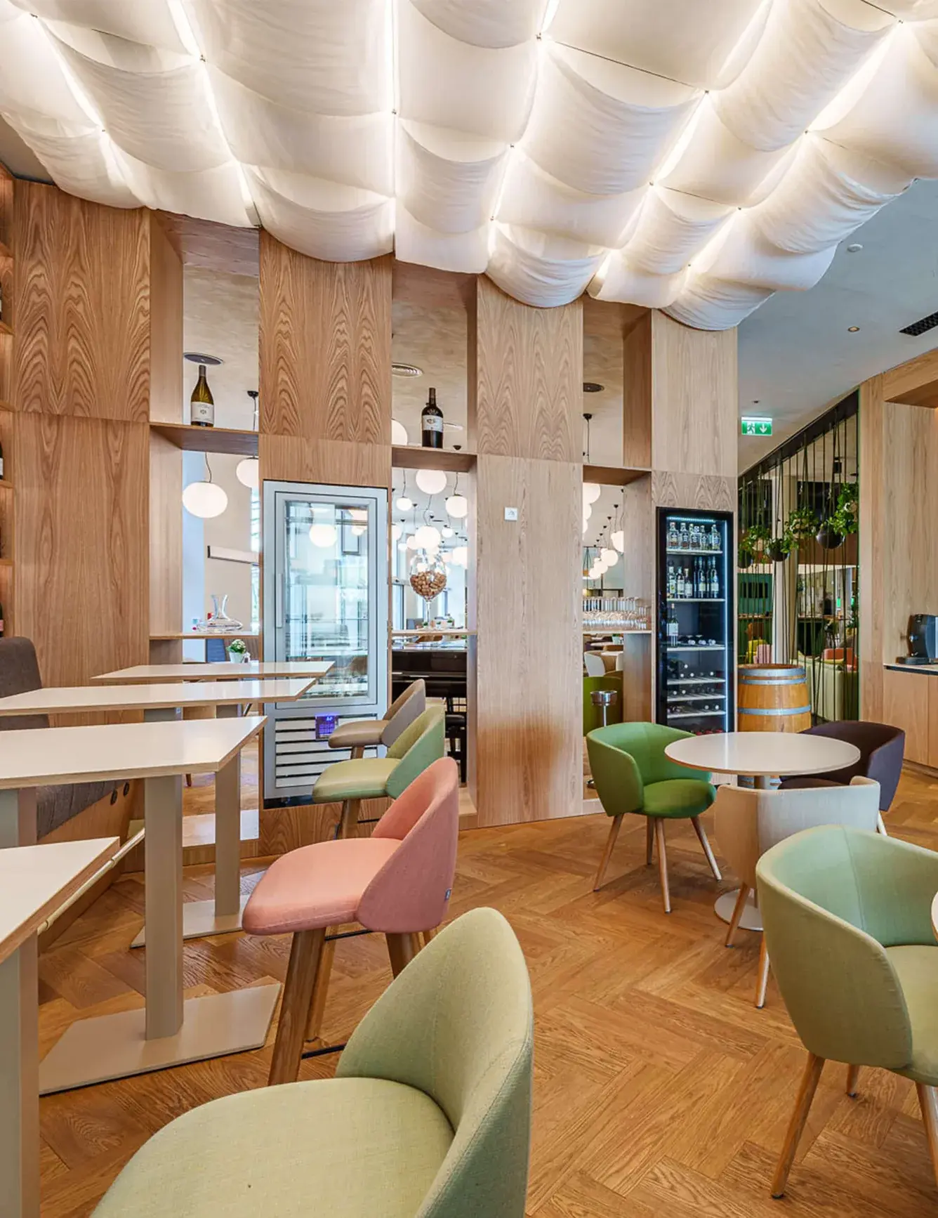 Pohled do moderního lobby hotelu Holiday Inn v Praze, kterému dominuje prosvětlený zvlněný podhled a dřevěná stěna s vinotékou, doplněná o barevná křesílka a dubovou podlahu Oakcent ve vzoru heringbone