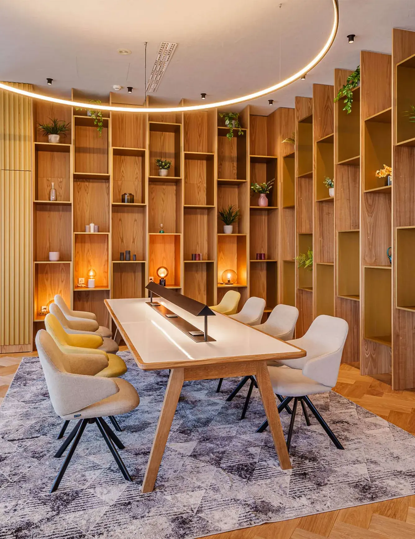 Interiér moderní zóny pro hosty v hotelovém lobby Holiday Inn s vysokou dřevěnou knihovnou sahající až ke stropu, která je doplněna o designové osvětlení a velký pracovní stůl s šedými a žlutými křesílky na dubové podlaze vzoru rybí kost.