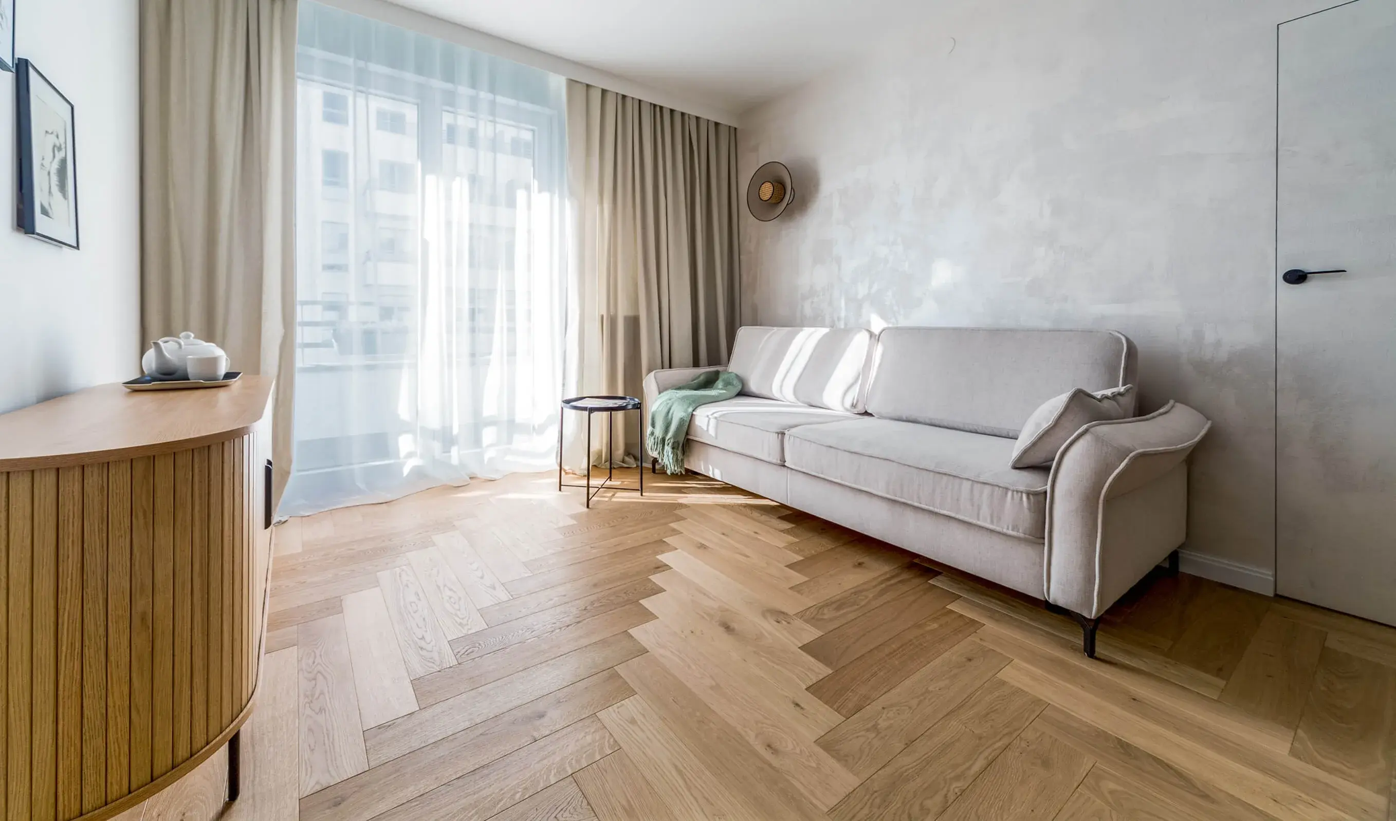 Prosvětlený interiér obývacího pokoje v mini apartmánu ve Varšavě, kterému dominuje rustikální dubová podlaha Oakcent Rustic položená ve vzoru stromečku (Herringbone), doplněná elegantní zaoblenou komodou a minimalistickou pohovkou.