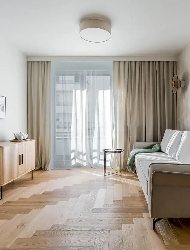 Prosvětlený obývací pokoj mini apartmánu ve Varšavě s dubovou podlahou Oakcent Rustic položenou do vzoru stromečku (Herringbone), doplněný minimalistickou pohovkou, dřevěnou komodou a jemnými závěsy.
