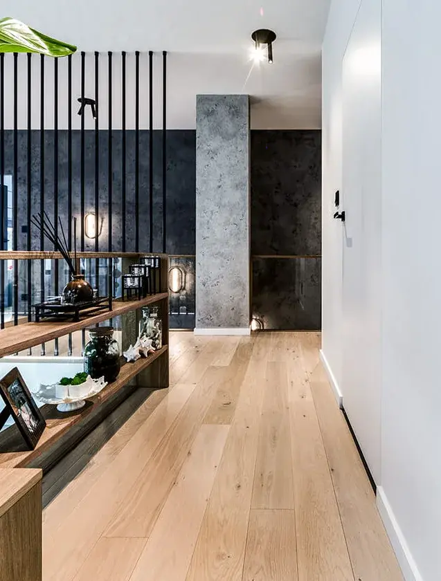 Detailní pohled na horní chodbu soukromé rezidence u Vratislavi s dřevěnou podlahou Oakcent Rustic v olejované úpravě Milky, kterou doplňuje designová policová sestava s dekoracemi a industriální dělící stěna z černých kovových tyčí na pozadí tmavé texturované stěny.