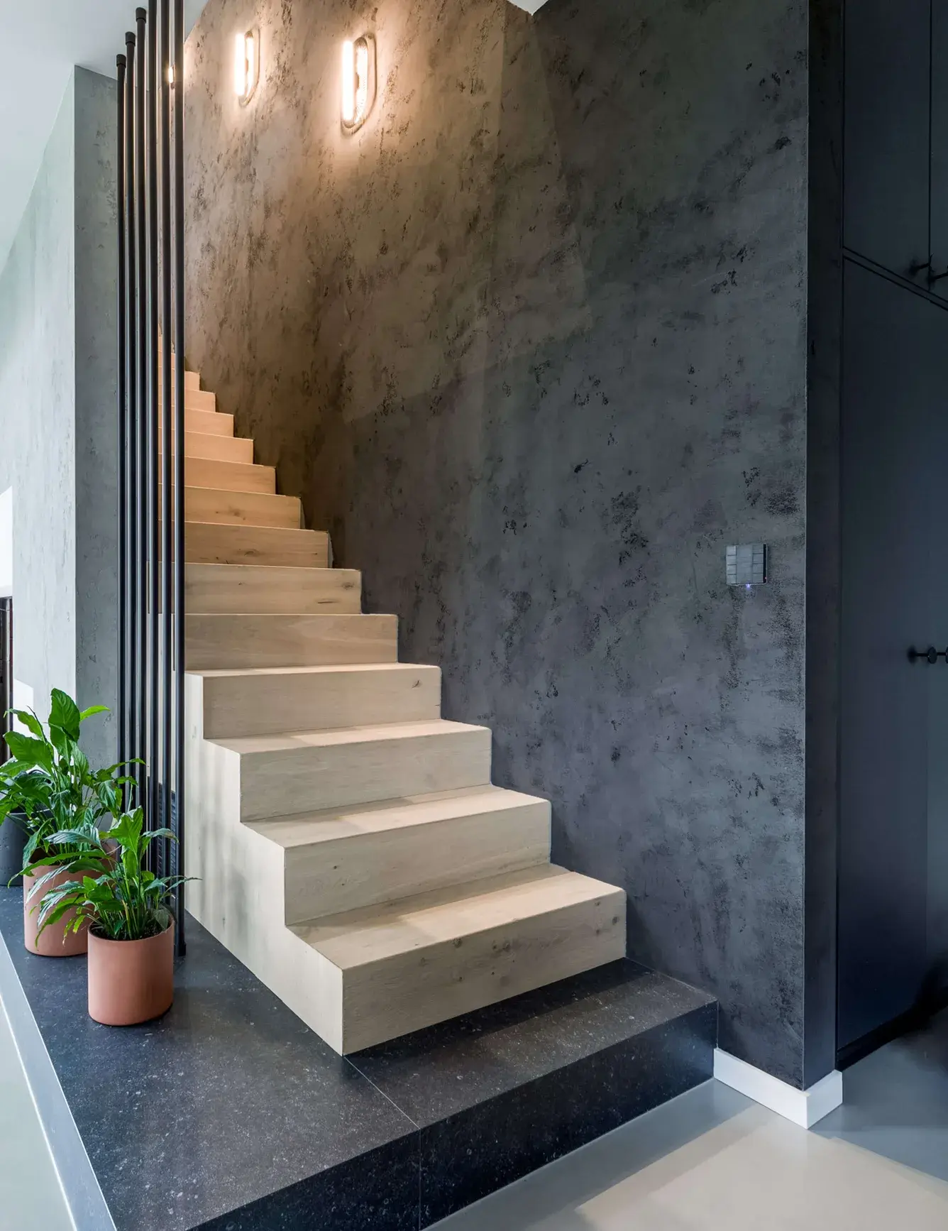 Moderní samonosné dřevěné schodiště v soukromé rezidenci u Vratislavi, vyrobené z dubu v odstínu Milky, které barevně kontrastuje s tmavou dekorativní stěrkou na stěně a černým kamenným soklem, doplněné o industriální kovové zábradlí a pokojové rostliny v terakotových květináčích.