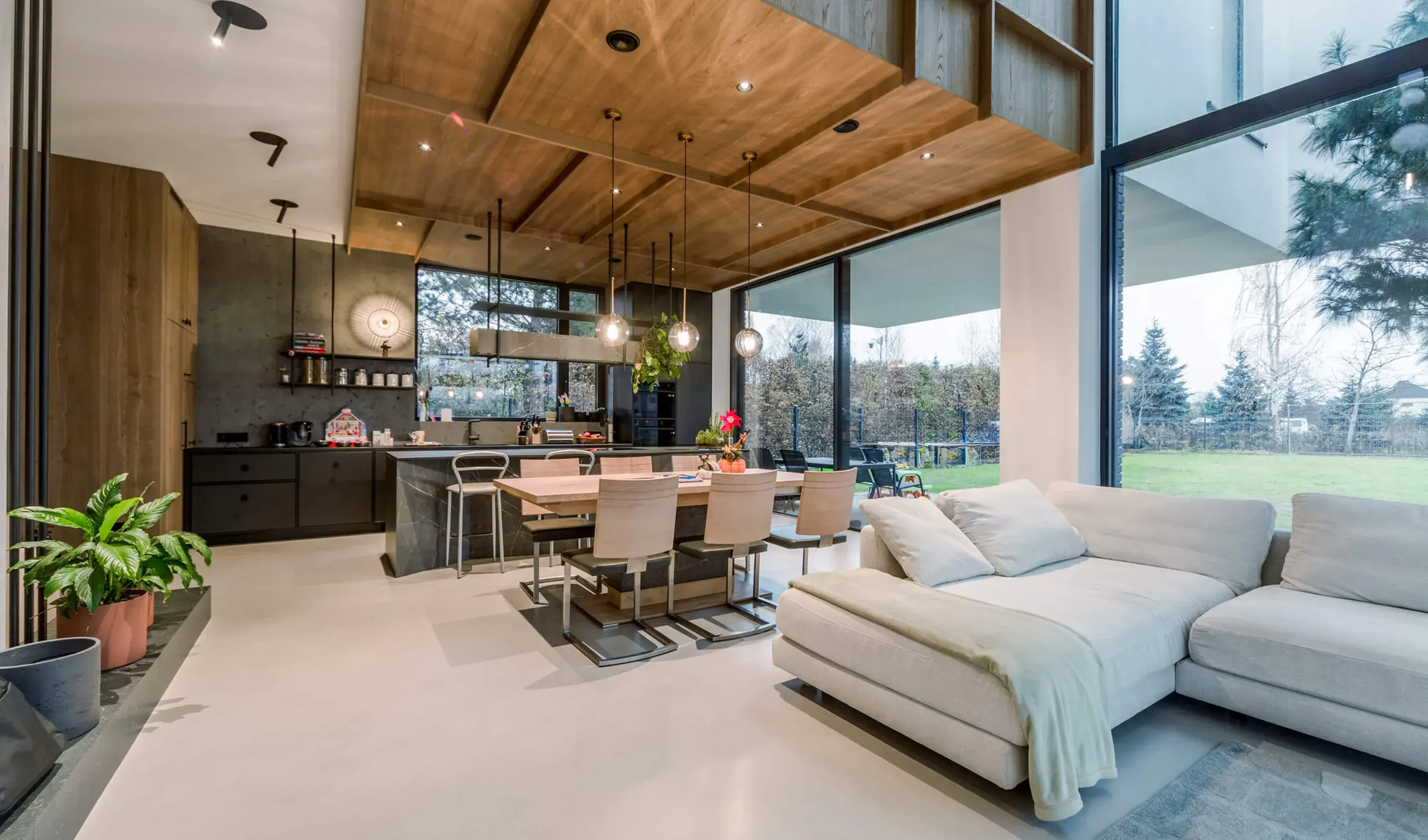 Moderní prostorný interiér soukromé rezidence u Vratislavi s dřevěným kazetovým stropem a světlou litou podlahou, kterému dominuje tmavý mramorový kuchyňský ostrůvek, minimalistická sedací souprava a jídelní stůl z masivního dubu před velkoformátovým oknem do zahrady.
