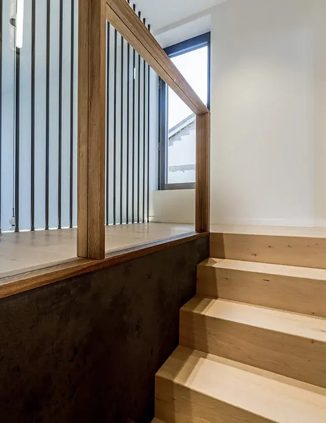 Pohled z dřevěného schodiště na galerii v patře rezidence u Vratislavi s dubovou podlahou Oakcent Rustic Milky, lemovanou proskleným zábradlím s masivním madlem a s průhledem na vertikální černé kovové tyče dělící stěny.