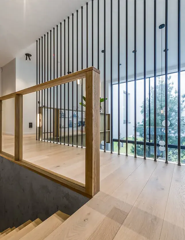 Pohled z horního patra rezidence u Vratislavi na galerii s dubovou podlahou Oakcent Rustic v odstínu Milky, kterou ohraničuje prosklené zábradlí s dřevěným madlem a výrazná dělící stěna z černých kovových prvků, s průhledem do spodního patra a k velkoformátovému prosklení.