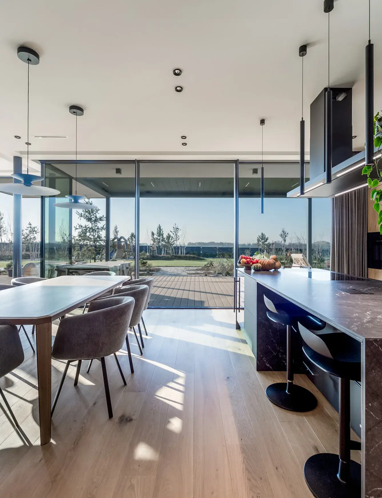 Pohled z moderní kuchyně s tmavým mramorovým ostrůvkem do prosvětlené jídelny a na zahradu, kde přírodní dubová prkna Oakcent Rustic v odstínu Pure s kartáčovaným povrchem a OML tmelem harmonicky propojují interiér s venkovní terasou.