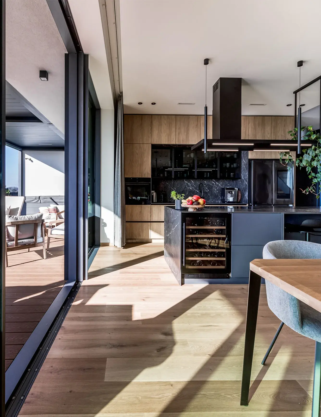 Pohled z moderní kuchyně s tmavým mramorovým ostrůvkem a vinotékou do prosvětleného interiéru domu v Tyniec Mały, kde přírodní dubová prkna Oakcent Rustic v odstínu Pure plynule navazují na venkovní terasu a ladí s dřevěným dekorem kuchyňských skříněk.