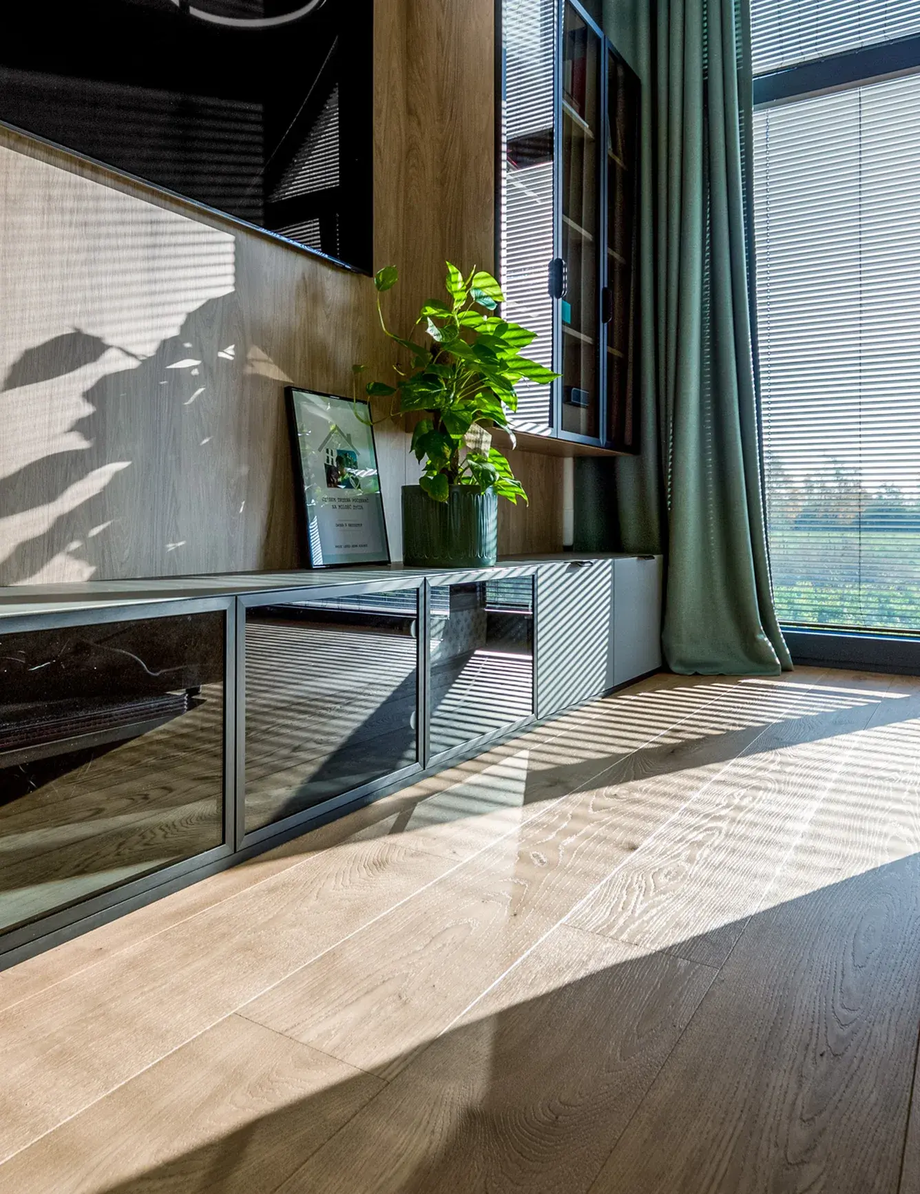 Detail obývací stěny v domě Tyniec Mały, kde sluneční paprsky pronikající přes žaluzie zdůrazňují kartáčovanou texturu dubové podlahy Oakcent Rustic v přírodním odstínu Pure.