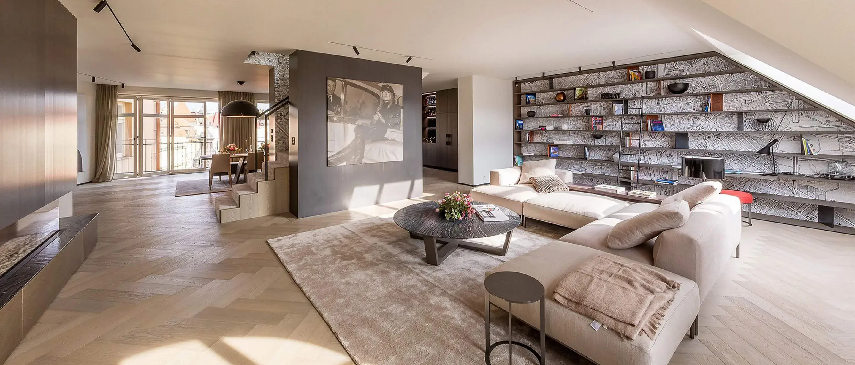 Luxusní interiér mezonetového Soukromého bytu v Praze s dubovou podlahou položenou ve vzoru maďarského stromu (Chevron) v odstínu Smoke, který doplňuje prostorná rohová sedací souprava, designová knihovna přes celou stěnu a moderní schodiště.