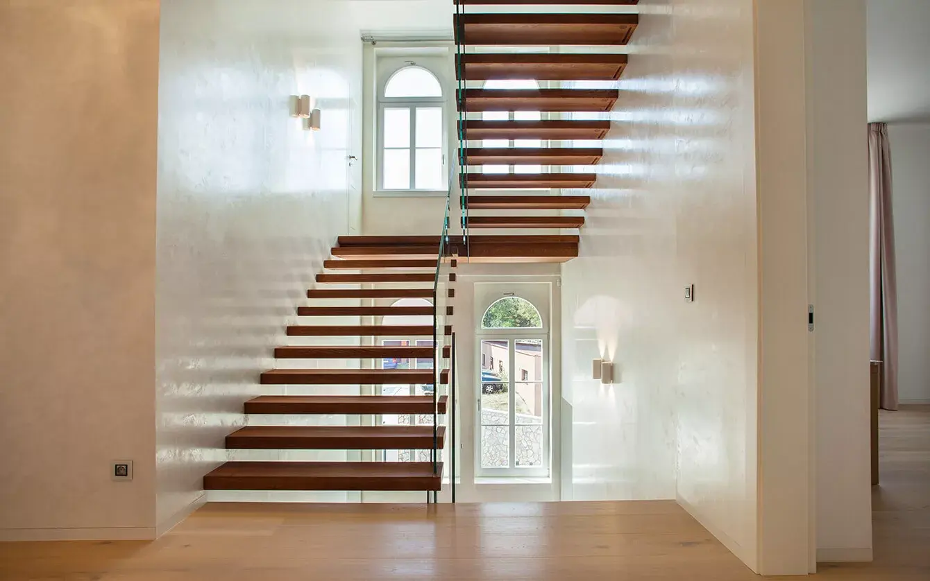 Interiér Vily Blaženka s moderním samonosným dřevěným schodištěm s proskleným zábradlím, které propojuje patra se světlou dubovou podlahou a je osvětleno přirozeným světlem z vysokých oken s půlkruhovým zakončením.