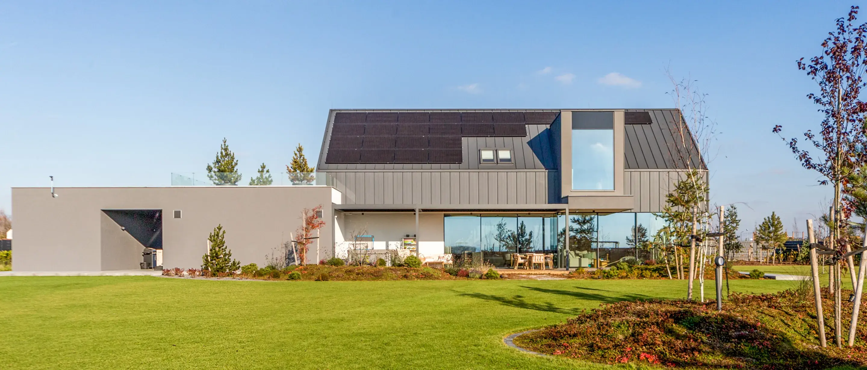 Exteriér moderního nízkoenergetického Soukromého domu Tyniec Mały se sedlovou střechou osazenou solárními panely, velkoformátovým prosklením v přízemí a upravenou zahradou s trávníkem.